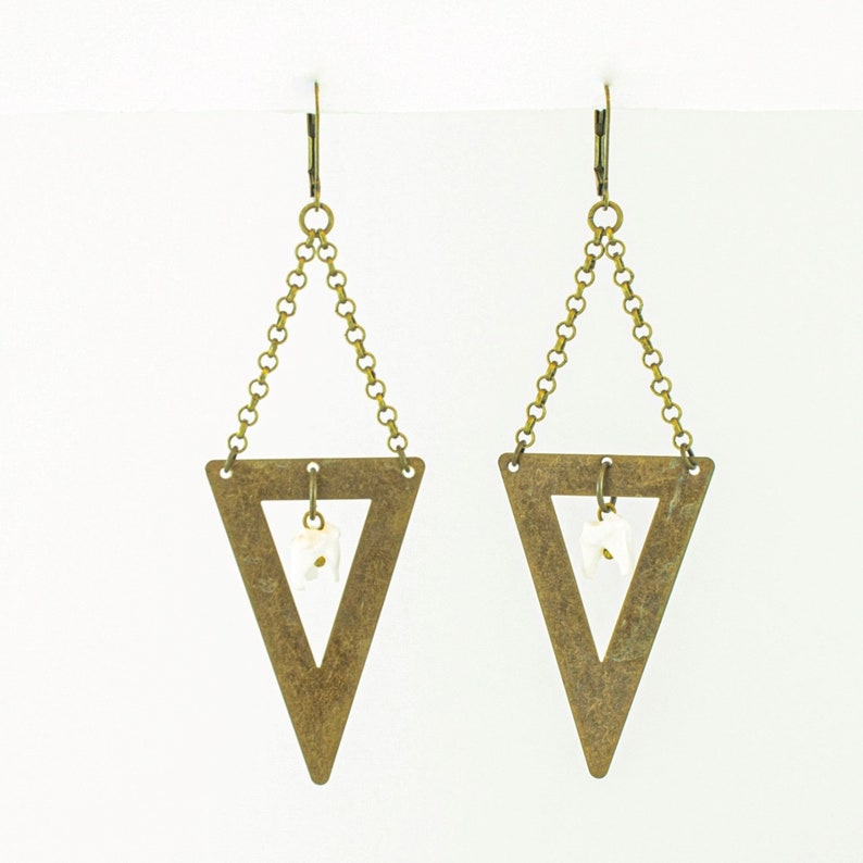 May include: A pair of brass-toned triangle earrings. Each earring features a large, open triangle pendant suspended from a chain and a small, white, irregular-shaped stone. The earrings have a simple, geometric design.