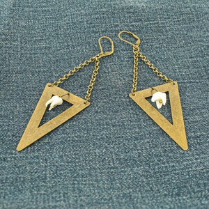 May include: A pair of brass-toned triangle earrings with a small white tooth charm inside each triangle. The earrings hang from a chain and have a lever-back closure. The earrings are displayed on a blue denim background.