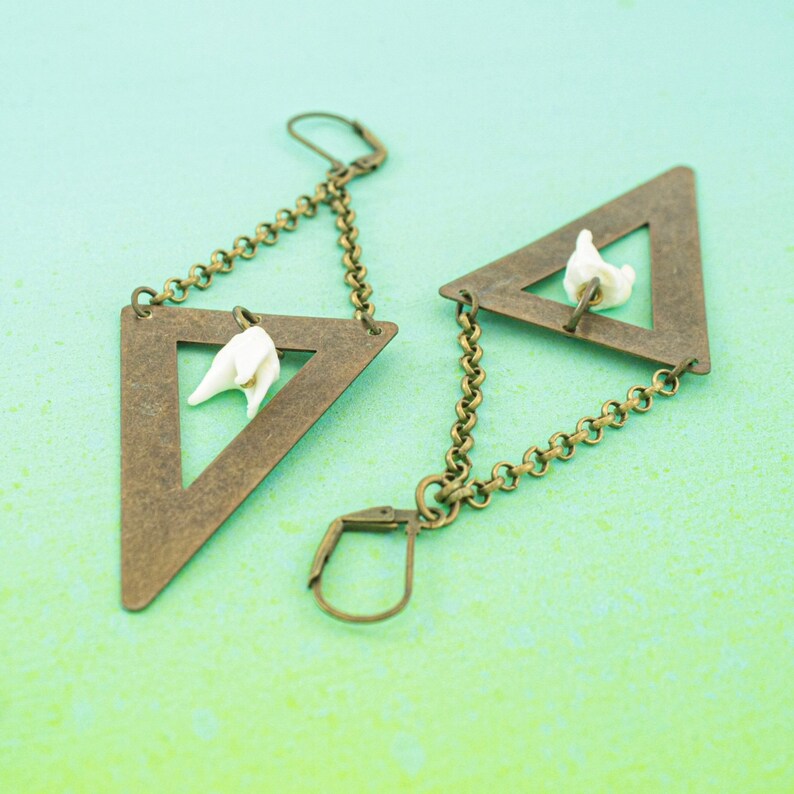 May include: A pair of unique earrings featuring bronze-colored triangular frames with a white floral accent. The earrings hang from a chain and have a lever-back closure. The design is set against a light teal background.