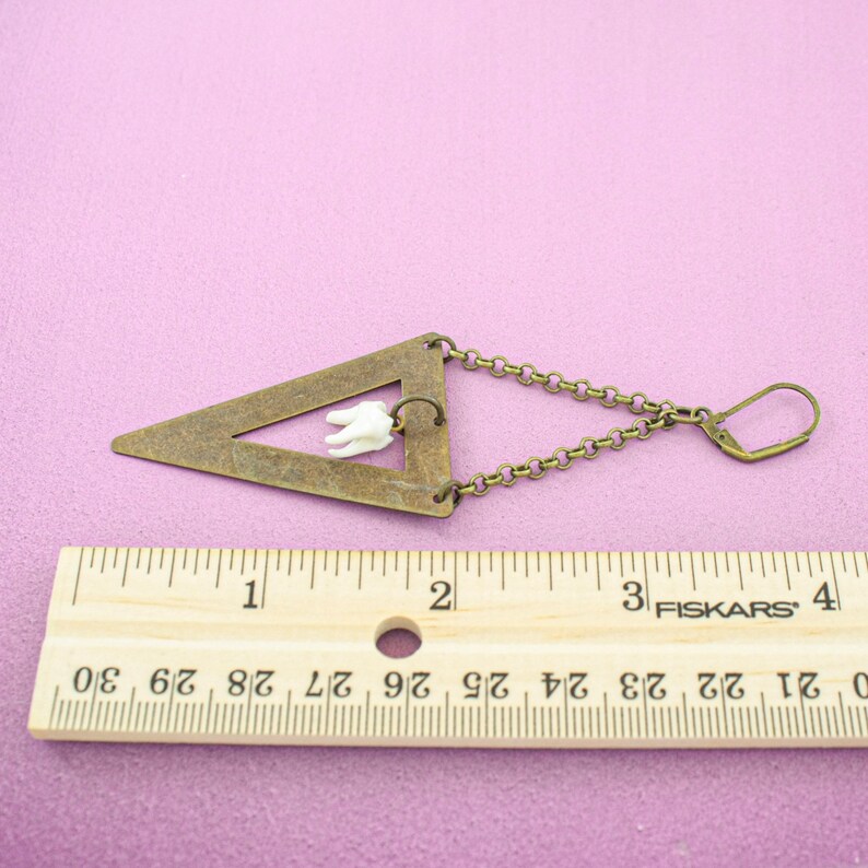 May include: A unique earring featuring a bronze-colored triangle with a small white tooth charm. The earring is connected to a chain and a hook closure. The ruler next to the earring shows measurements in inches.