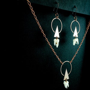 Puede incluir: Un collar y pendientes de cobre con un colgante de tri&aacute;ngulo geom&eacute;trico y un dije de diente. El collar tiene una cadena delicada y un colgante circular con un dije de tri&aacute;ngulo.