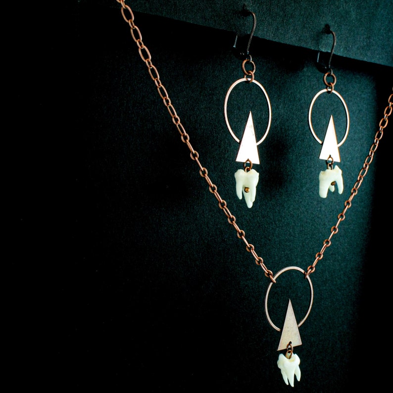 Puede incluir: Un collar y pendientes de cobre con un colgante de tri&aacute;ngulo geom&eacute;trico y un dije de diente. El collar tiene una cadena delicada y un colgante circular con un dije de tri&aacute;ngulo.