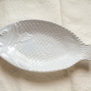 Peut inclure: Plat de service en céramique blanche en forme de poisson avec une surface texturée ressemblant à des écailles.