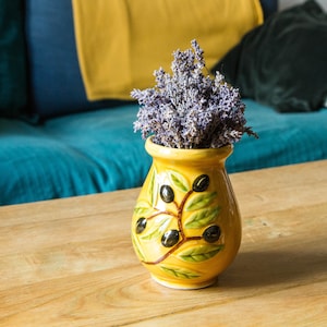 Peut inclure: Un vase en céramique jaune avec un motif de branche d'olivier vert contient un bouquet de fleurs de lavande séchées. Le vase est posé sur une table en bois.