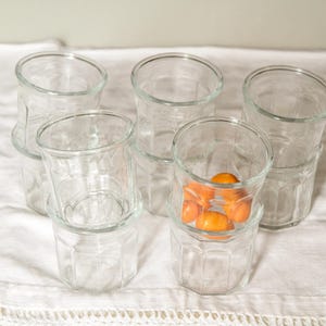 May include: Several clear glass tumblers with a faceted design, some stacked. One tumbler contains small, orange fruits. The glasses are arranged on a white, textured surface. The glasses are suitable for serving drinks.