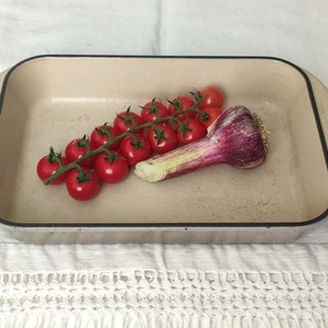 Könnte beinhalten: Eine weiße Keramik-Backform mit schwarzem Rand, die einen Zweig rote Kirschtomaten und eine einzelne violette Knoblauchknolle enthält.