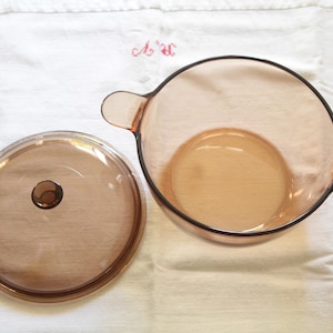 Peut inclure: Une cocotte ronde en verre ambré dotée de deux poignées latérales et d'un couvercle circulaire assorti avec un petit bouton central. Cet ustensile de cuisine transparent présente une finition lisse et polie, vue de dessus.