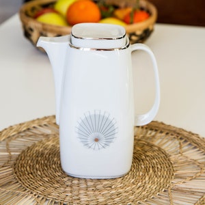 Può includere: Caffettiera in ceramica bianca con bordo argentato e un design grigio e argento sul davanti. La caffettiera è appoggiata su un sottopentola in vimini intrecciato.