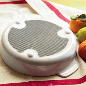 Può includere: Piatto da forno in ceramica bianca con un inserto a rete. Il piatto è appoggiato su una tovaglia a righe rosse e bianche con tre arance e un limone.