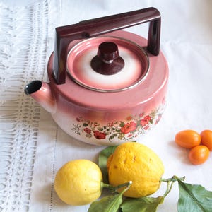 Peut inclure: Une théière vintage en émail rose et blanc avec une poignée et un couvercle marron foncé. La théière présente un motif floral avec des roses rouges et des feuilles vertes. Deux citrons et trois petites oranges sont disposés près de la théière sur une nappe blanche.