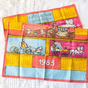 Puede incluir: Dos paños de cocina de lino vintage con un diseño de calendario de 1983. Los paños presentan ilustraciones de carruajes tirados por caballos en varios colores, incluyendo rosa, amarillo y azul. El texto "Les Transports à Travers les âges" es visible.