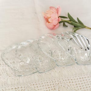 Op de afbeelding: Vier heldere glazen schelpvormige schalen zijn gerangschikt op een wit kanten tafelkleed. Een roze pioenroos en groene bladeren zijn op de achtergrond. De schalen hebben een gestructureerd, geribbeld ontwerp.