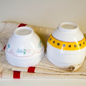 May include: Two white ceramic bowls with floral and geometric designs. The bowls are stacked on top of each other and are sitting on a beige and red striped towel.