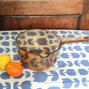 Op de afbeelding: Een amberkleurige glazen steelpan met een handvat, geplaatst op een blauw-wit gebloemd tafelkleed. Naast de steelpan liggen een gele citroen en een oranje mandarijn. De achtergrond is een houten kast.