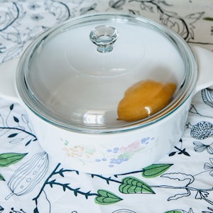 May include: A white ceramic casserole dish with a glass lid. The dish has a floral pattern and is sitting on a white tablecloth with a black floral design.