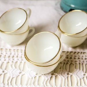 Puede incluir: Cuatro tazas de té de vidrio de leche blanco con ribete dorado apiladas en pares sobre un mantel de crochet blanco.