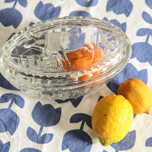 Pode incluir: Um prato oval de vidro transparente com uma cena detalhada de fazenda na tampa, contendo itens laranja. Dois limões amarelos repousam ao lado do prato em um tecido estampado floral azul e branco.