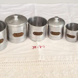 Puede incluir: Conjunto de seis botes de aluminio vintage con tapas. Cada bote tiene una etiqueta marrón con una palabra. Los botes están dispuestos en fila sobre un mantel blanco. El mantel tiene un borde decorativo.