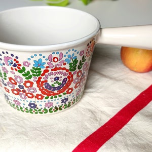 Könnte beinhalten: Weißer Keramiktopf mit einem Blumendesign in den Farben Rosa, Blau, Grün und Rot. Der Topf hat einen einzigen Griff und steht auf einer weiß-rot gestreiften Oberfläche.