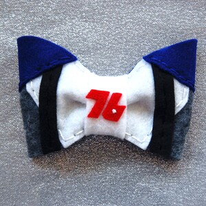 May include: A white felt bow tie with a red felt number "76" in the center. The bow tie has blue and gray felt accents.