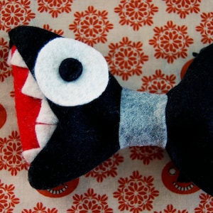 May include: A black felt bow tie with a white eye and red teeth, resembling a shark. The bow tie is on a white background with a red floral pattern.