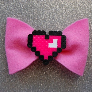May include: A pink felt bow with a pixelated pink heart in the center. The heart is outlined in black beads.