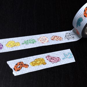 May include: Washi tape with a repeating pattern of colourful snake illustrations. The snakes are in various colours and patterns, including yellow, green, pink, and orange, on a light blue grid background. The tape is partially unrolled, showing the design.