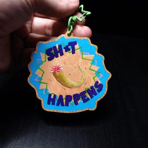 以下が含まれることがあります： 青とベージュのデザインの木製キーホルダー。「SH*T HAPPENS」の文字が青色で書かれています。ピンクの花が付いた緑色のピクルスがキーホルダーの中央にあります。緑色の星型のクリップが上部に取り付けられています。
