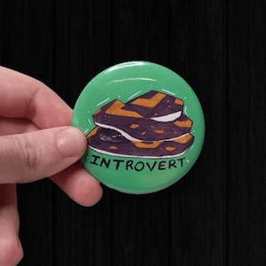 May include: A green badge with the word "INTROVERT" in white text. The badge features a graphic of a stack of brown and purple shapes. The badge is held in a hand against a dark background.