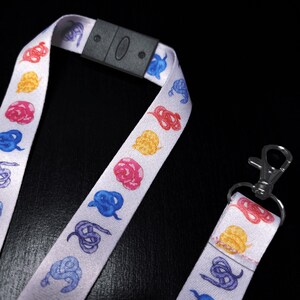 May include: White lanyard featuring a repeating pattern of colourful snake illustrations. The lanyard has a black plastic clasp and a silver metal clip. The snakes are in shades of red, yellow, blue, and pink.