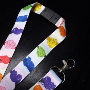 May include: A white lanyard featuring a repeating pattern of colourful, stylised flower illustrations. The flowers are in shades of red, pink, blue, green, and yellow. The lanyard has a black plastic clasp and a silver metal clip.
