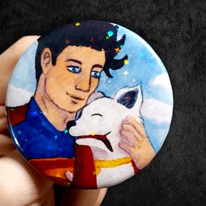 May include: A circular badge with a watercolour illustration of a man in a blue shirt and red cape embracing a white dog. The background is light blue with stars.