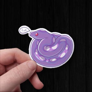 May include: A purple snake sticker with white markings and a red eye. The snake is coiled, with a speech bubble saying "fuck". The sticker is held by a hand against a black background.