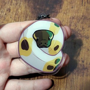 May include: A clear acrylic keyring featuring a cartoon illustration of a coiled snake with a white body, yellow patches, and dark brown spots. The snake has a dark brown head with black eyes and a small black nose, attached to a black metal ring.