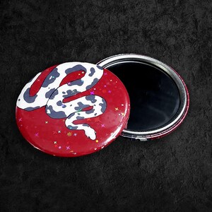May include: A red compact mirror featuring a white and black snake design. The mirror's cover is decorated with a stylized snake illustration and colourful star-shaped confetti. The open mirror reveals a reflective surface.