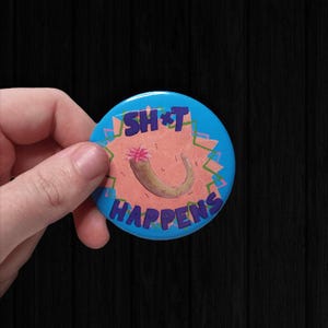 以下が含まれることがあります： 紫色の文字で「SH*T HAPPENS」と書かれた丸い青色のボタン。茶色の物体とピンクの花の漫画イラストが描かれています。ボタンは人が持っています。