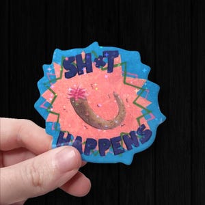 以下が含まれることがあります： 青いキラキラ文字で「SH*T HAPPENS」と書かれたステッカー。ステッカーの中央はピンクとオレンジで、茶色の曲線とピンクのとげのあるデザインがあります。ステッカーは星型のアクセントが付いた青い縁で囲まれています。