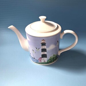 May include: White ceramic teapot with a lid, spout, and handle. The teapot features a coastal design with a lighthouse, seagulls, and a small house. The background is a light blue color.