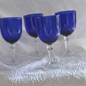 May include: A set of four cobalt blue glass goblets, each featuring a deep blue rounded bowl and a clear glass stem with a spherical accent at the top, resting on a circular clear glass base, creating a uniform and classic drinkware collection.