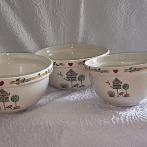 May include: Three cream-colored ceramic mixing bowls of varying sizes, each featuring a decorative rim with green leaves, small red hearts, and a central illustration of a birdhouse on a post surrounded by small trees and a flying bird.