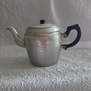 May include: A silver-toned metal teapot featuring a hammered texture on the body, a curved spout, and a black handle. The lid is topped with a small black knob, and a decorative beaded border encircles the upper section of the teapot body.
