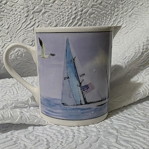 May include: A white ceramic pitcher featuring a nautical scene with a sailboat on blue water, a flying seagull, and an American flag on the sail. The pitcher has a curved handle and a pouring spout, with a smooth finish and a cylindrical body shape.