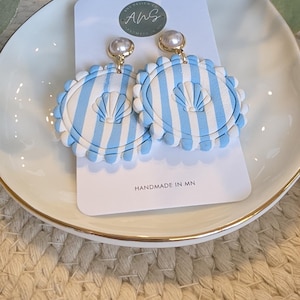 May include: A pair of handmade earrings with a seashell design. The earrings feature a light blue and white striped pattern with a pearl accent. The text "HANDMADE IN MN" is visible on the product card.