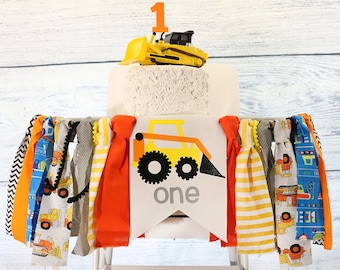 May include: A white cake with a yellow construction vehicle on top and a banner that says "one" with a yellow construction vehicle on it. The cake is decorated with a gray and white fabric garland.