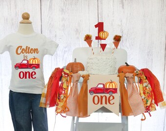 May include: A white onesie with the text "Colton is one" and a red truck with a pumpkin in the bed. A red and orange banner with a red truck and pumpkin reads "one". The banner is strung with orange and red fabric scraps.