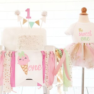 May include: A birthday party setup with a white cake, a banner, and a tutu outfit. The cake is topped with a pink number 1 and decorative flags. The banner features an ice cream cone graphic and the word "one". The outfit includes a white onesie with "the sweet one" text.