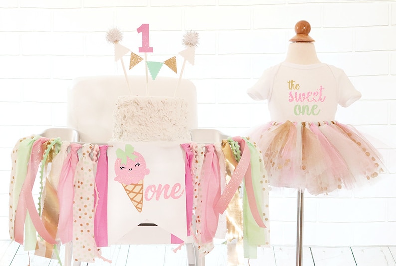 May include: A birthday party setup with a white cake, a banner, and a tutu outfit. The cake is topped with a pink number 1 and decorative flags. The banner features an ice cream cone graphic and the word "one". The outfit includes a white onesie with "the sweet one" text.
