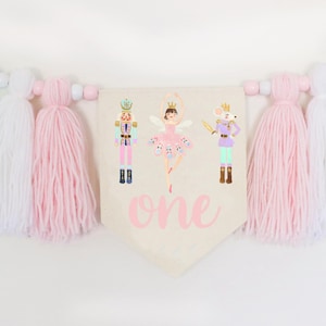 Puede incluir: Un banderín con temática de ballet, con un banderín central con ilustraciones de una bailarina, un cascanueces y un rey ratón. La palabra "one" está escrita en rosa. El banderín está adornado con borlas en verde menta, blanco y rosa, y cuentas de madera.