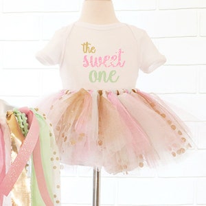 May include: A white baby bodysuit with the text "the sweet one" in gold, pink, and green lettering. The bodysuit is paired with a pink, gold, and white tutu. A decorative garland with pink, gold, and green ribbons is on the left.