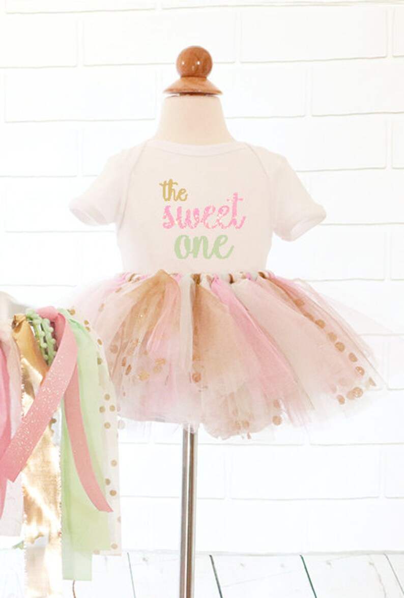May include: A white baby bodysuit with the text "the sweet one" in gold, pink, and green lettering. The bodysuit is paired with a pink, gold, and white tutu. A decorative garland with pink, gold, and green ribbons is on the left.